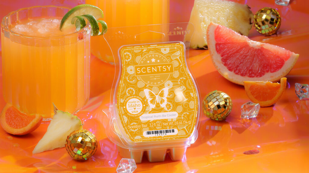 Scentsy's Tropical Rum-Ba Cooler Wax Bar sits with it's clamshell lid open on an orange countertop. Gold disco balls, pitchers full of fruit juice, and grapefruit and pineapple slices surround the wax bar  amidst gleaming ice cubes.