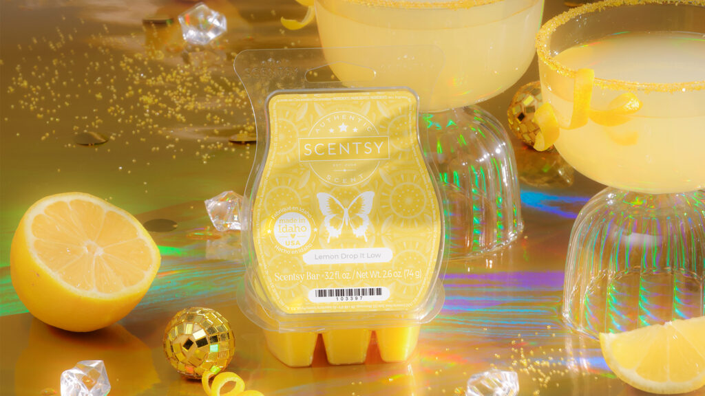 Scentsy's Lemon Drop it Low Wax Bar sits on a shimmering gold tabletop with an open, yellow clamshell lid. The Wax bar is surrounded by lemon wedges, gold disco ball party decor, and yellow-colored cocktails in tall glasses.