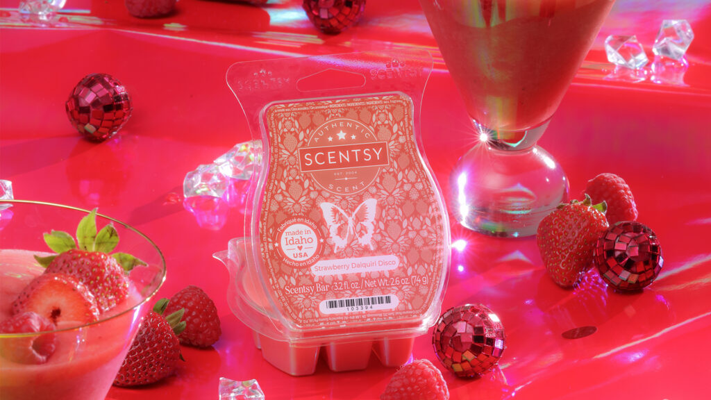 Scentsy's Strawberry Daiquiri Disco Wax Bar sits with it's red, clamshell lid open on a shimmering red tabletop. Redbright red strawberries and glittering red disco ball party favors decorate the tabletop, along with shimmering ice cubes and frosty strawberry cocktails in tall, cone-shaped glasses. 