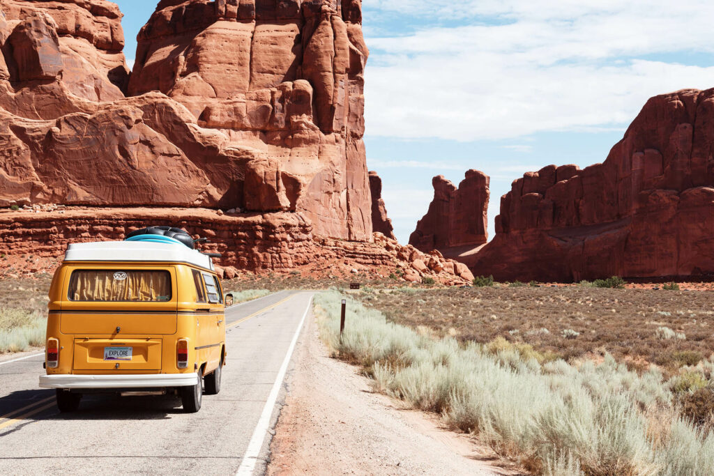 Yellow van driving on dry desert highway with giant red rock structures and a brush field 