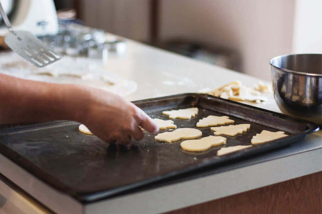 Person placing cookie cutouts on a baking sheet while a mess awaits in the background