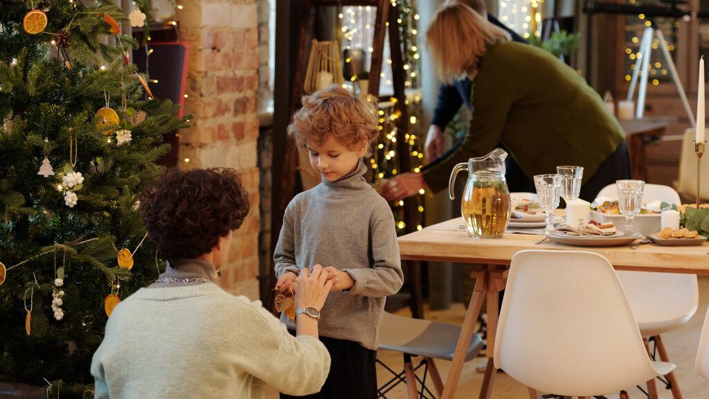 A little boy being handed an ornament by an adult male to hang up on the tree beside a table set for holiday feast with christmas decor all around