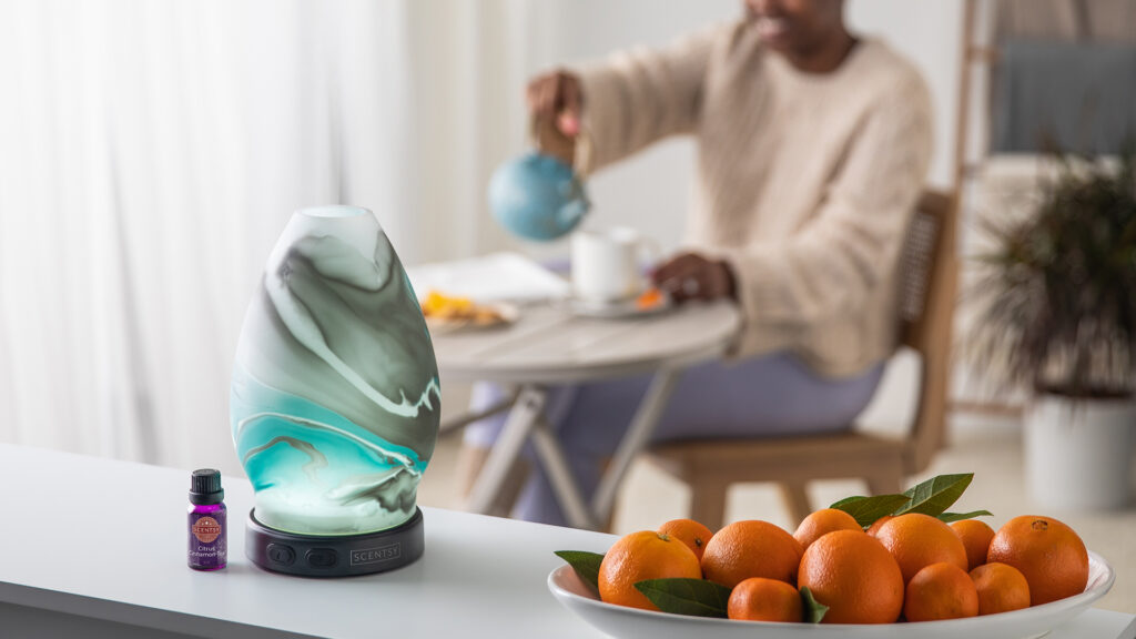 A scentsy oil diffuser diffusing citrus cinnamon tea scented essential oils beside a fruit bowl of oranges with a woman in the background pouring herself a cup of tea at the dining table