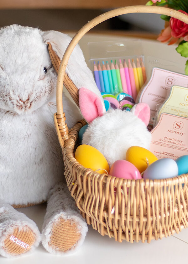 A Bailey the Bunny Scentsy Buddy rests on a table near an Easter basket filled with plastic eggs, wax melt Scentsy Bars and other products and gifts.