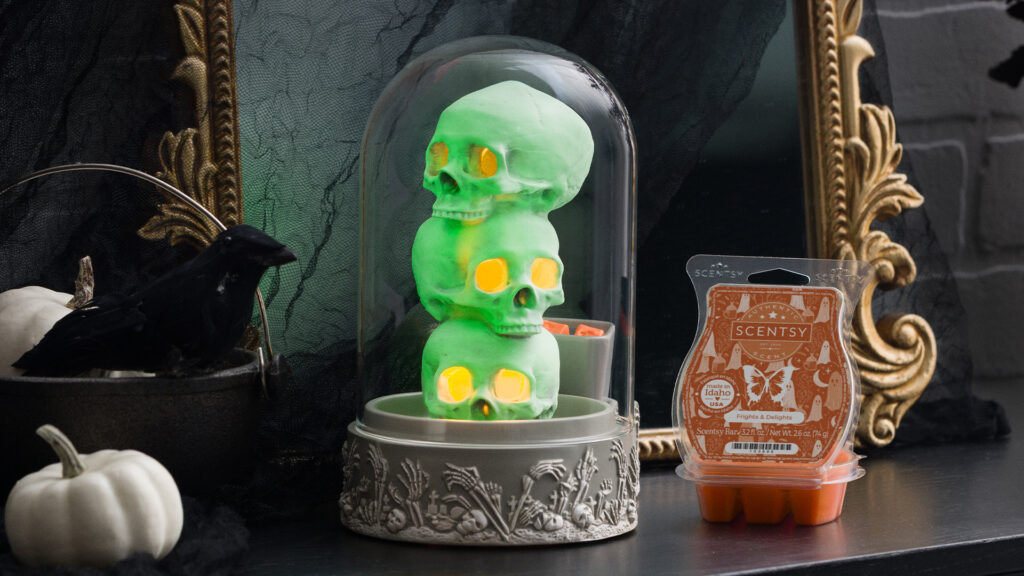 Encased in a glass cloche, the Forsaken Tower Wax Warmer depicts three stacked skulls bathed in a glow-in-the-dark finish and with yellow light glowing from the eye-sockets. The warmer is displayed in a spooky scene on black shelf beside the Frights & Delights wax bar, pale white pumpkins, a stuffed crow, and a gilt-framed mirror.