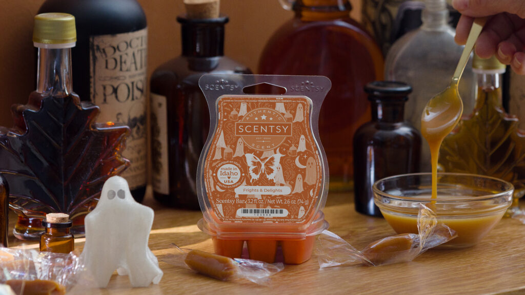 The orange-labled Frights & Delights Scentsy Wax Bar sits on a table full of vials and bottles. A toy ghost, caramel candies, and a spoon stirring melted carmel in a glass bowl surround the Scentsy wax melt. 