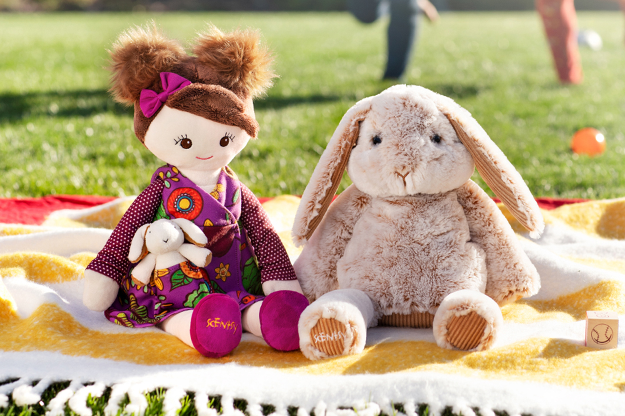 Scentsy Buddies on a blanket outside with people playing in the background