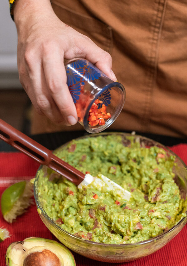 Person making guacamole with ingredients surrounding the bowl