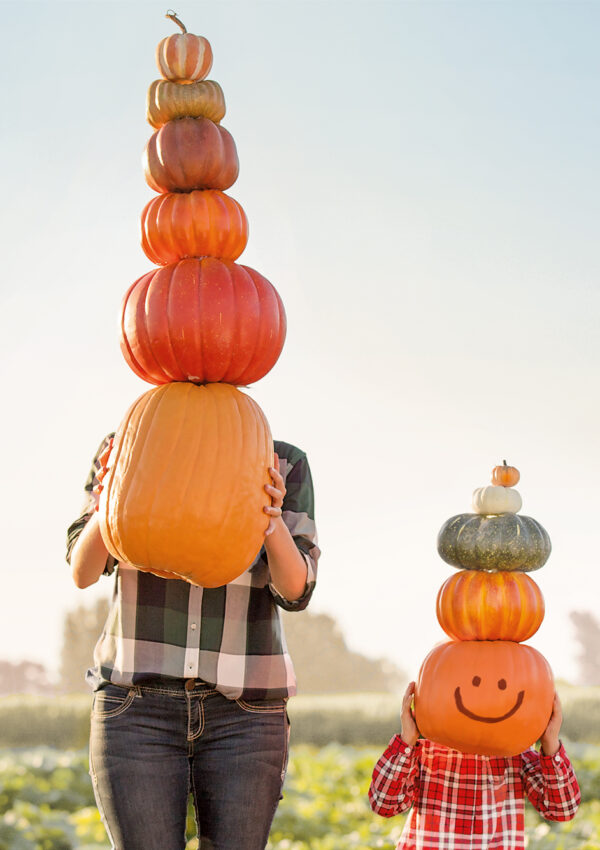 Adult and child holding giant stacks of pumpkins up in-front of their faces