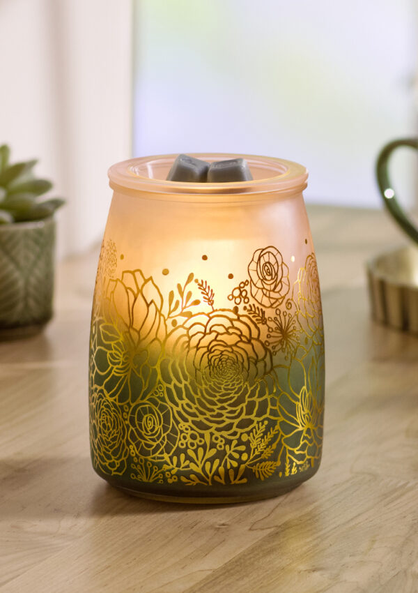 Succulents and house plants in green, grey, and brown pots sit on a wooden table. In the back, out of focus, is a green coffee cup. Front and center, the Scentsy Tranquil Glow Warmer glows yellow. Gold-etched flowers emboss it's green and white gradient design, and four golden wax melt cubes sit in the warming dish on top.