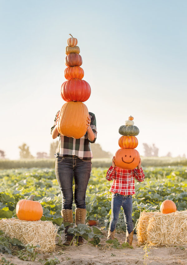 A mother and her daughter standing in the middle of a pumpkin patch. The mother holding a tall stack of 6 pumpkins with the biggest on the bottom and the daughter with 5 stacked pumpkins, the biggest on the bottom with a black smiley face drawn on it