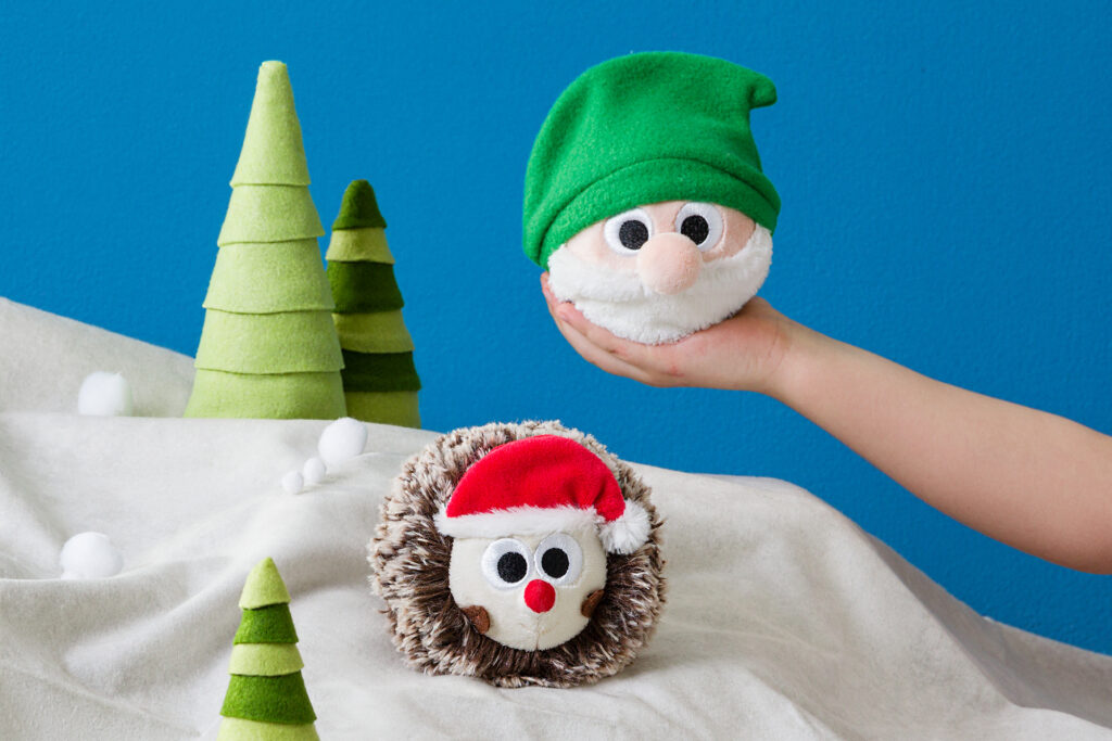 The Gnome and Holiday Hedgehog Scentsy Bitty Buddy stuffed animals sitting in front of a cartoon-like snowy forest backdrop