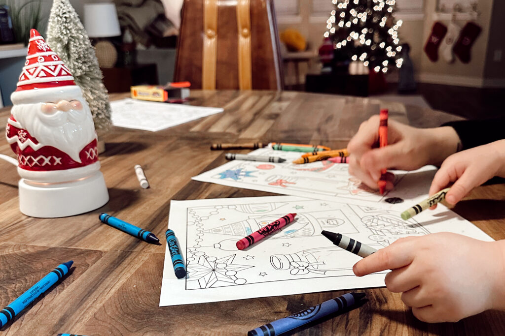 Kids coloring Scentsy coloring pages on the table with crayons scattered, a Santa Claus wax warmer and a lit Christmas tree and stockings in the background