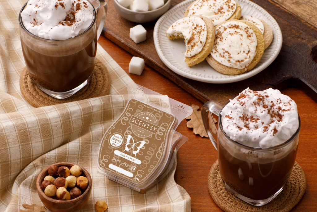 Scentsy wax melts scented in hug in a mug sitting on a dining table beside a bowl of hazelnuts, cups of hot cocoa, sugar cubes and plate of cookies