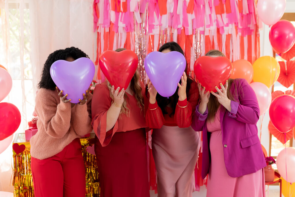 Four girl friends celebrating Galentine's Day together taking a photo with heart shaped balloons held up in front of their faces in front of a valentines day inspired back drop