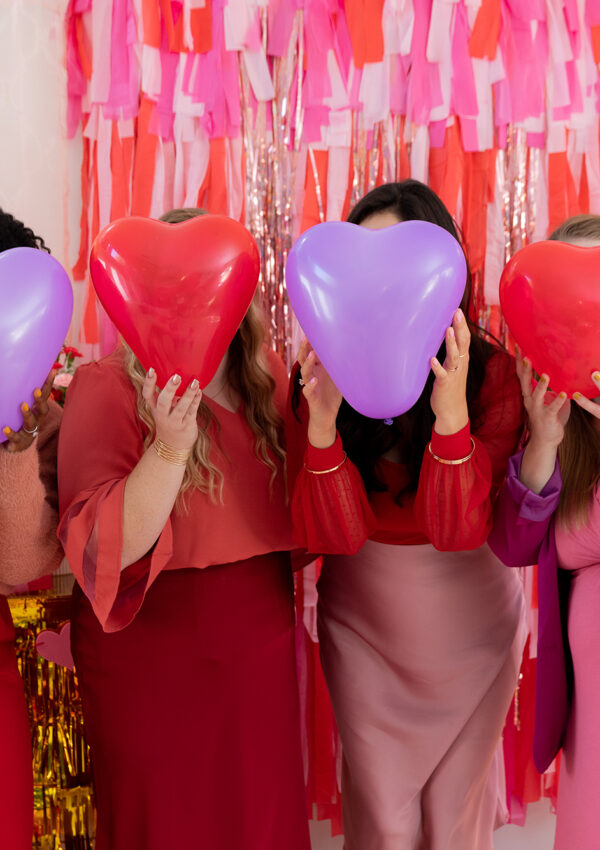 Four girl friends celebrating Galentine's Day together taking a photo with heart shaped balloons held up in front of their faces in front of a valentines day inspired back drop
