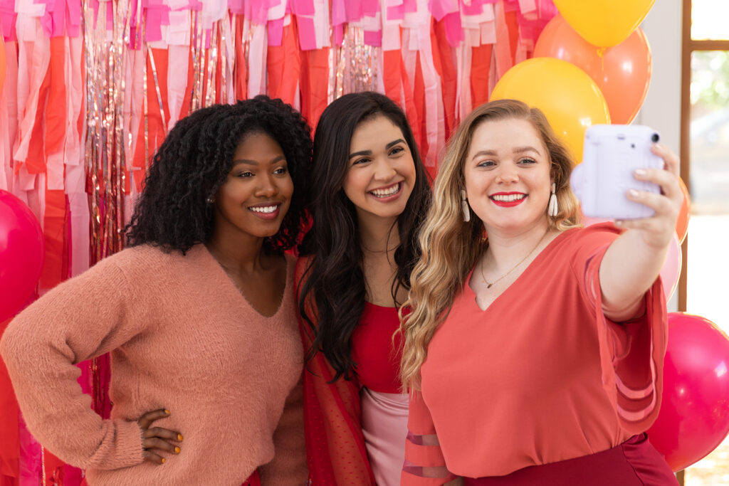 Three friends in front of a Galentine's Day Party decorated backdrop taking a selfie and capturing memories