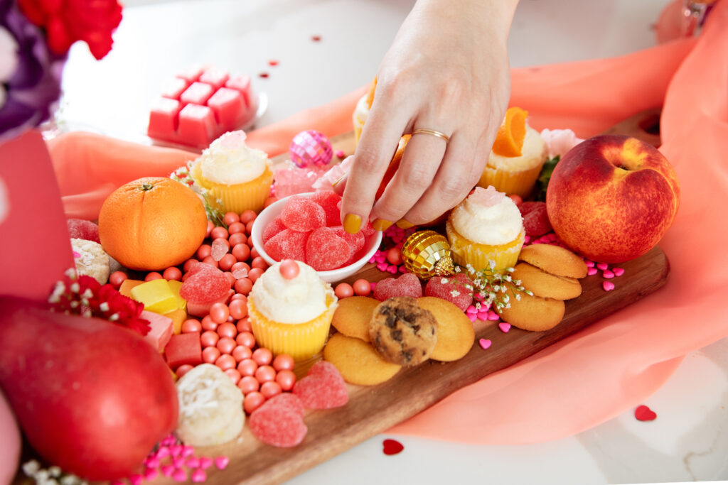A charcuterie board with candies, cupcakes and sweet fruit like clementines and peaches all festive for a Galentine's Day party