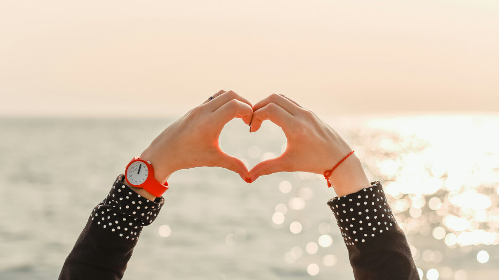 Hands held up making the shape of a heart with an ocean sunset in the background.