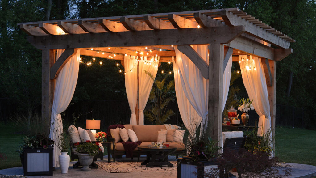 A backyard gazebo lit up with string lights with patio furniture, a drink station, and plants