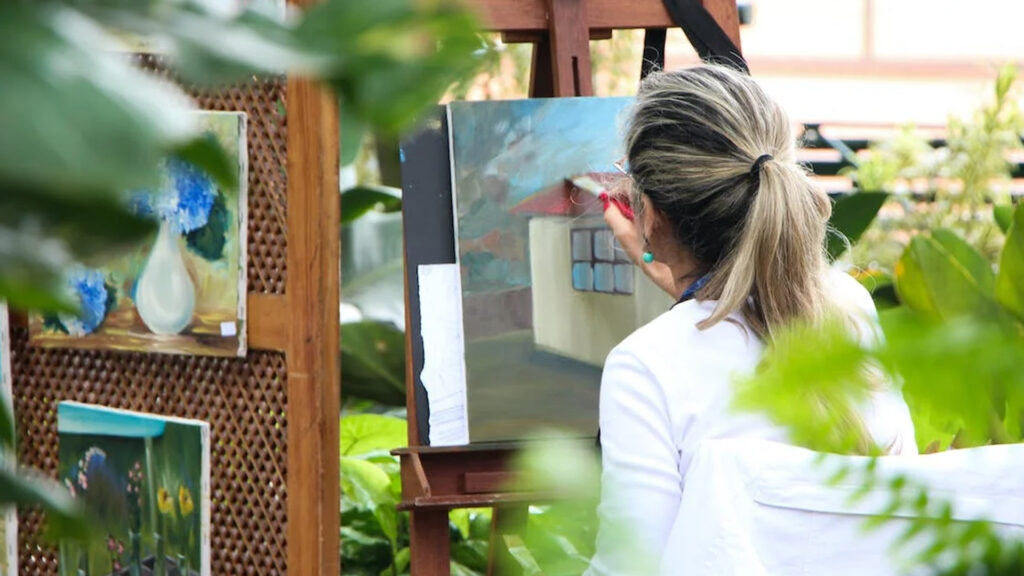 A woman painting outside on her patio a little cream house with a red roof