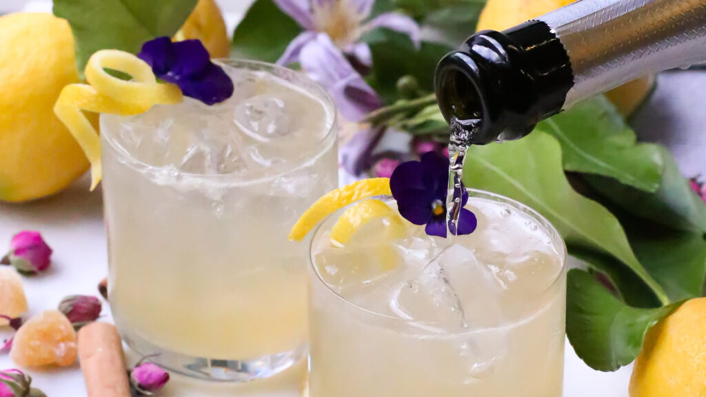 A beverage with an edible purple flower and lemon peel arrangement with prosecco being poured in