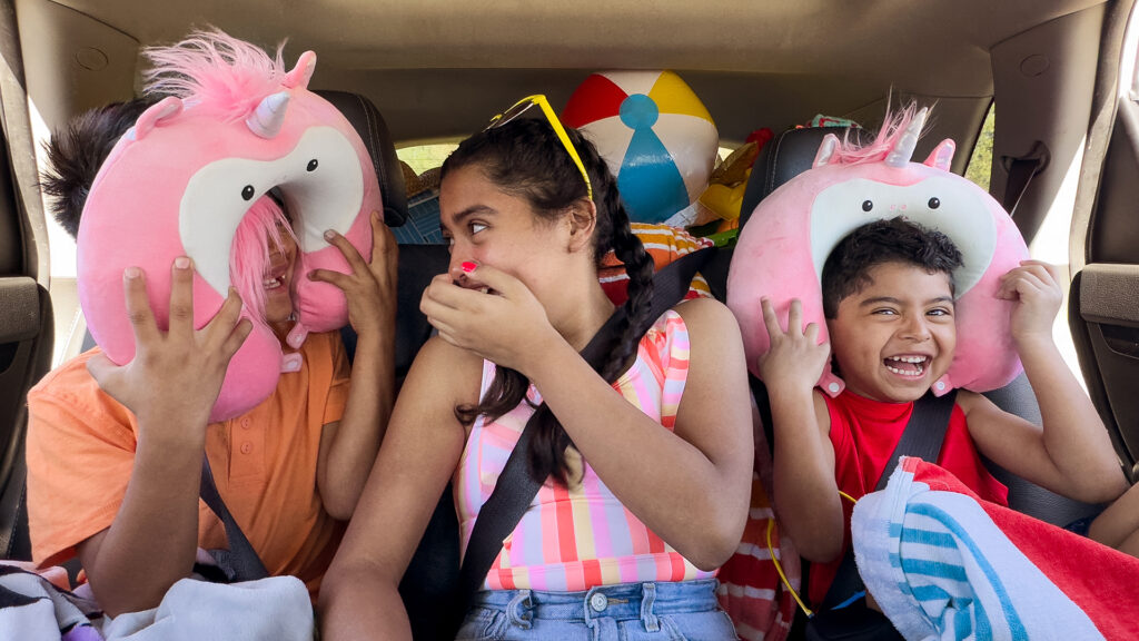 three kids sitting in the car on a road trip using the unicorn Scentsy buddy travel neck pillow