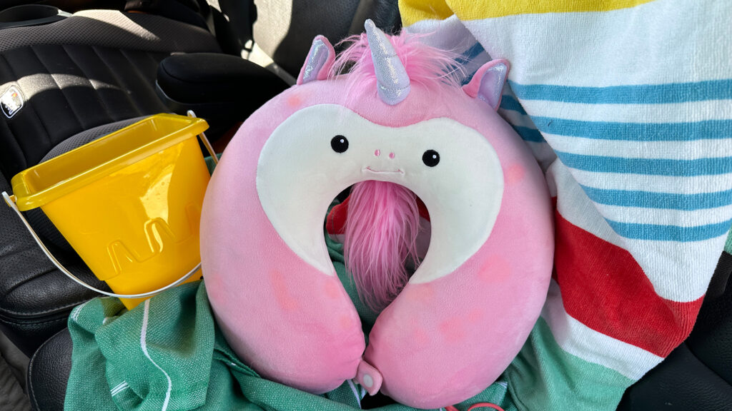 The Unicorn Scentsy Buddy travel pillow  sitting in the car beside a car seat, sand bucket and beach towel