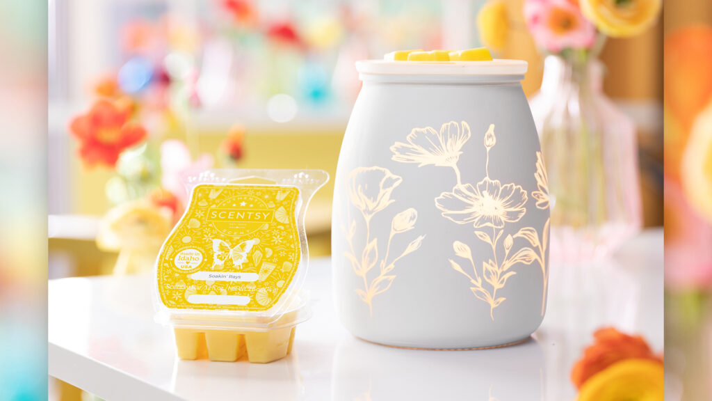Scentsy's Flower Garden wax warmer and a Soakin' Rays scented wax bar sitting on a white table with a vase of flowers blurred in the background