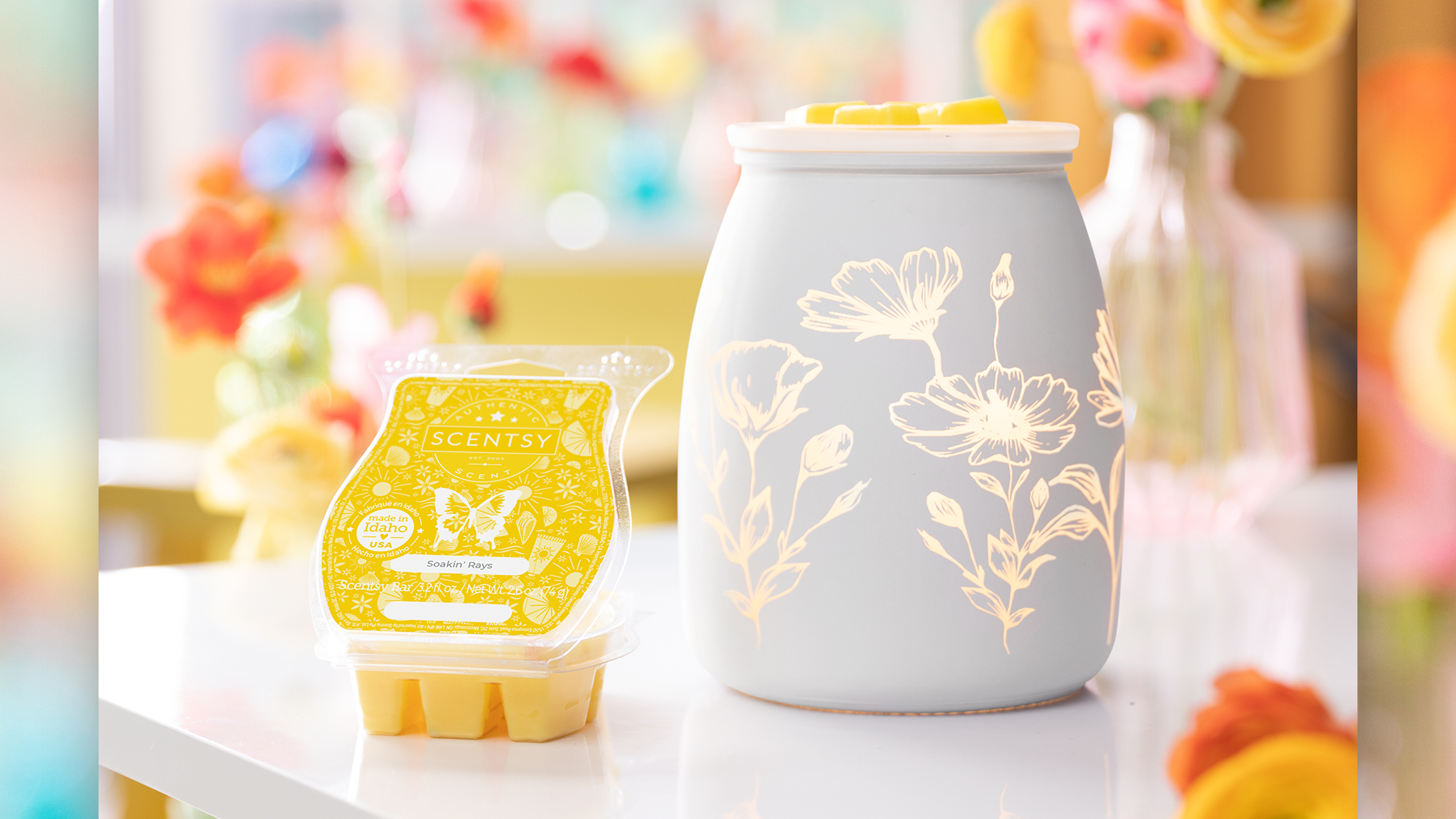 Scentsy's Flower Garden wax warmer and a Soakin' Rays scented wax bar sitting on a white table with a vase of flowers blurred in the background