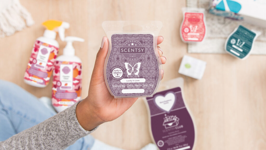 A woman's hand holding a Lucky in Love scented wax bar with scentsy cleaning spray, cotton cleanups, a scentsy wax brick and other wax bars laying on the floor