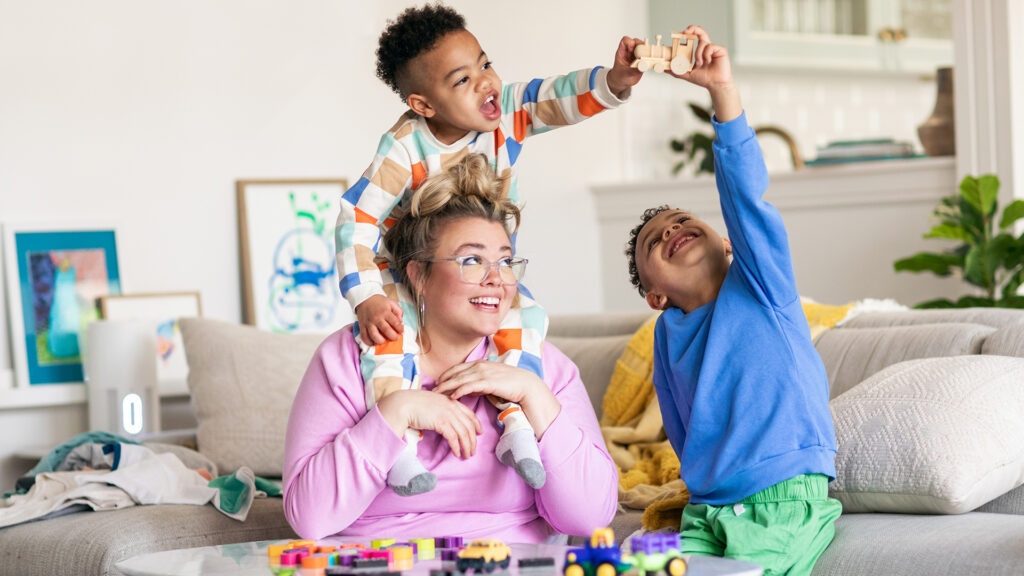 A woman and her two sons sitting in the living room on the couch playing with wooden train toys with a scentsy air purifier in the background on a side table