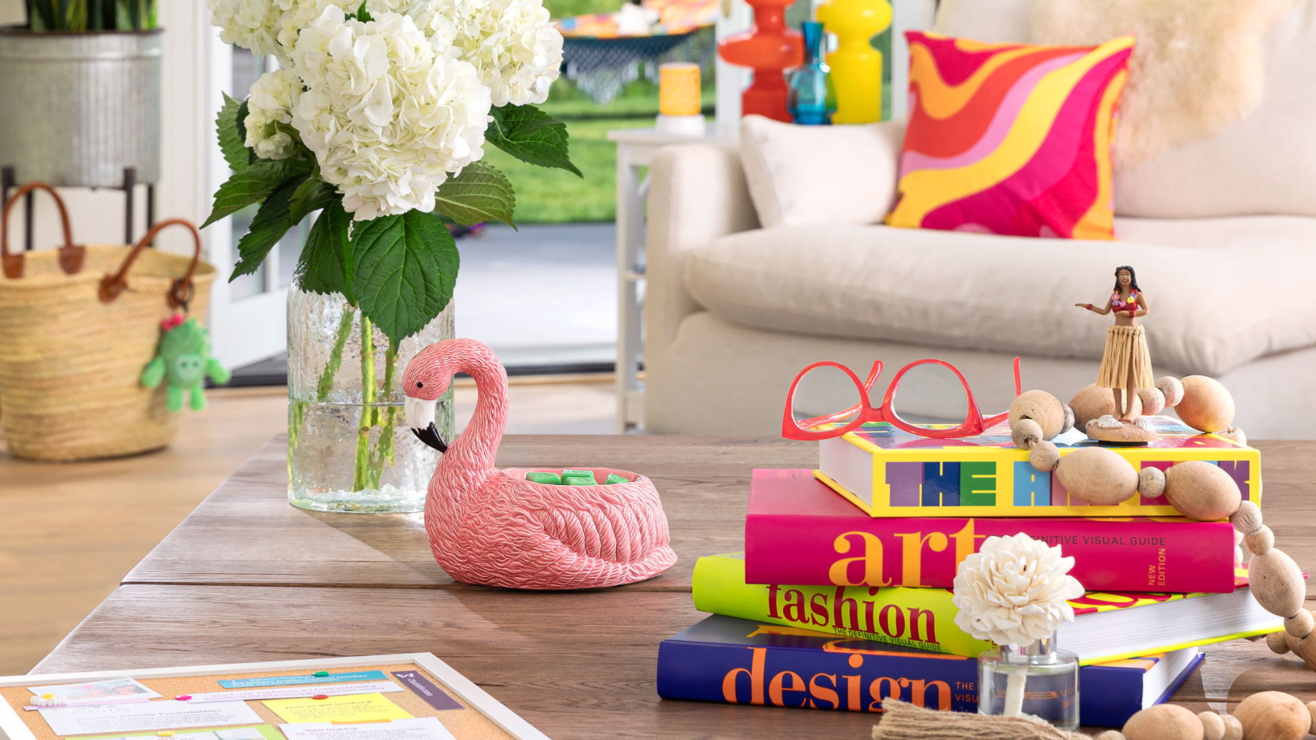 Living room full of vibrant summer colors and on coffee table sits a pink flamingo wax warmer, fragrance flower, stack of books, reading glasses, a hula girl and vase with white flowers in it.