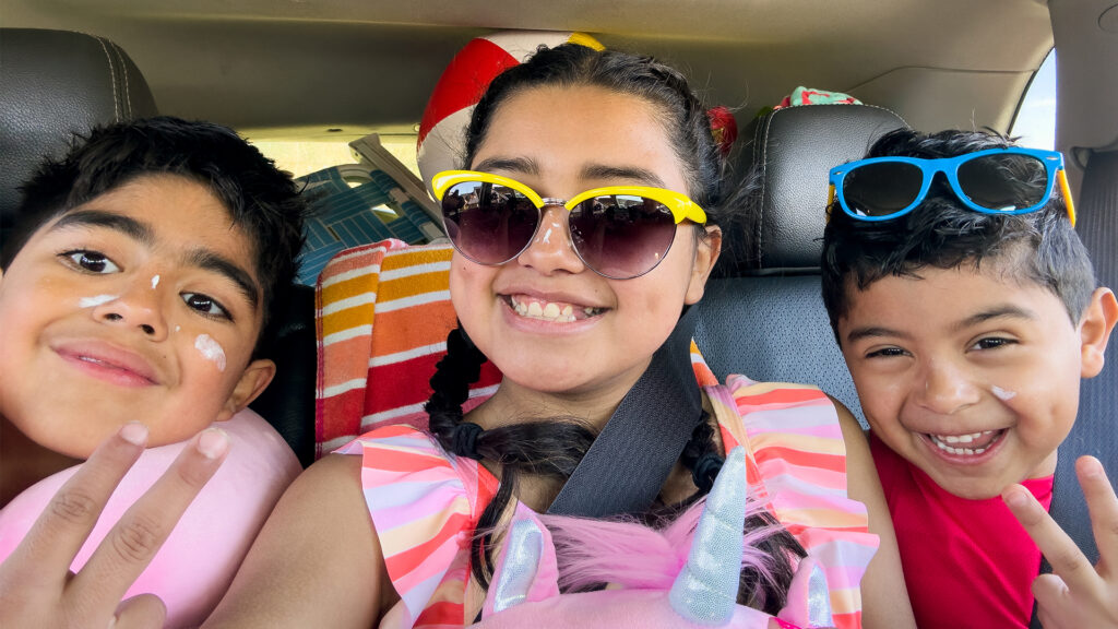 three kids in the car taking a selfie in their sunglasses and swimsuits with sunscreen on and the trunk full of beach chairs and other items