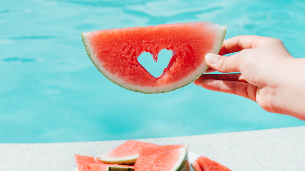 A person sitting by a pool holding up a piece of watermelon with a little heart shape cut out