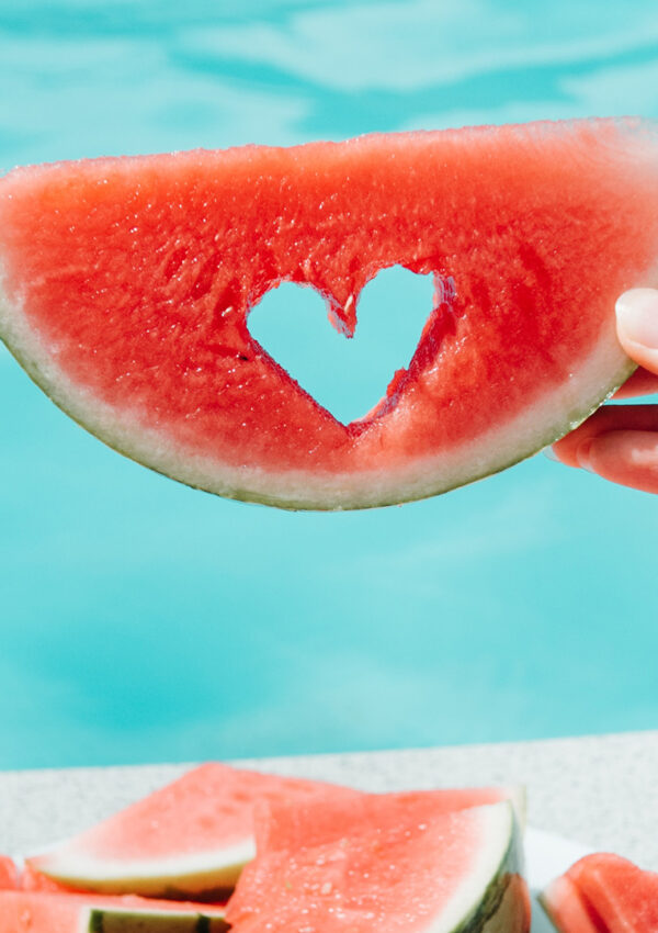 A person sitting by a pool holding up a piece of watermelon with a little heart shape cut out