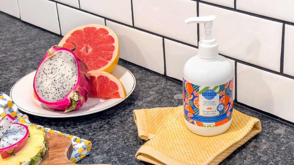 Tangerine and sugarcane scented hand soap sitting on a kitchen counter on a yellow dish towel beside a plate full of grapefruit, dragon fruit and pineapple