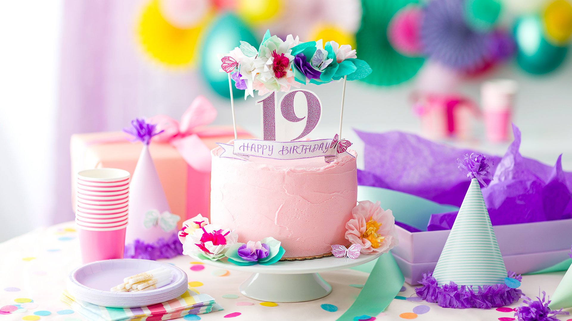 A table full of Birthday party décor with a pink cake that has the numbers 19 and a happy birthday floral sign with a butterfly to celebrate scentsy's 19th birthday