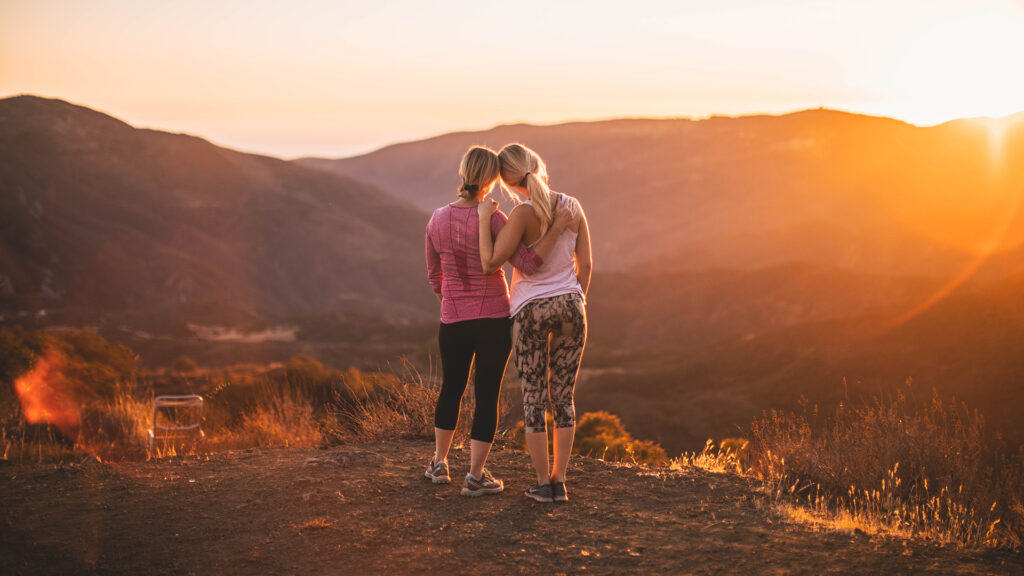 Two girls on a hike standing on the top of a mountain with their arms around each other watching the sunset