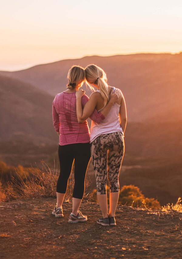 Two girls on a hike standing on the top of a mountain with their arms around each other watching the sunset