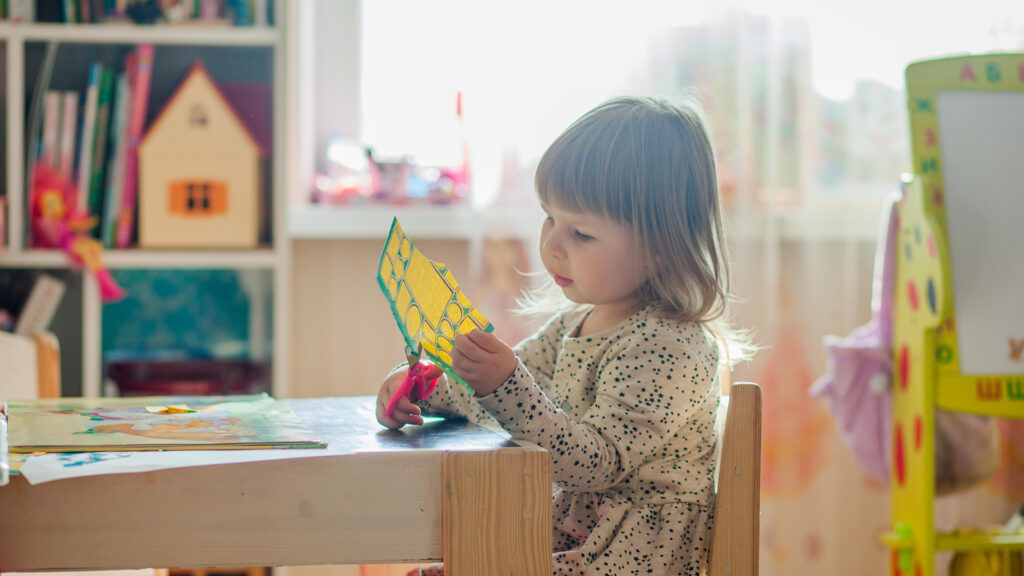 A little girl sitting in at her desk in a classroom cutting a paper with pink scissors