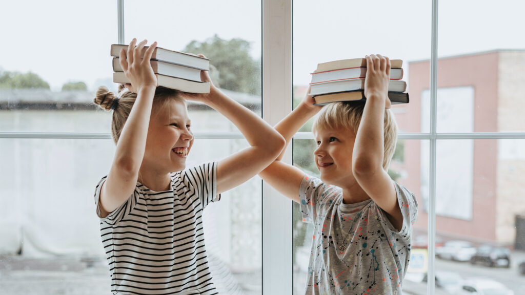 A young boy and girl laughing and balancing stacks of books on their heads