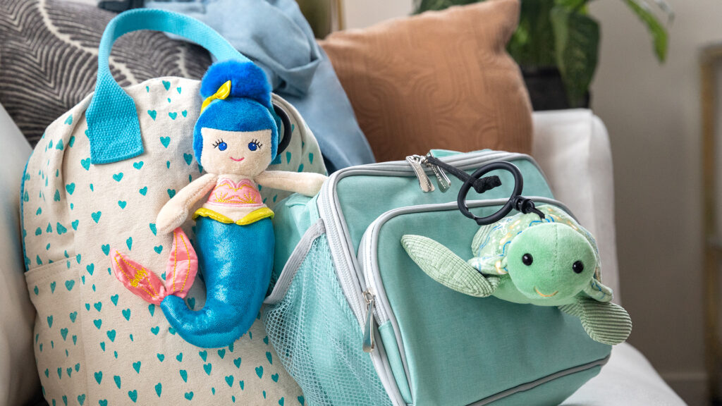 A white backpack with little blue hearts and a blue lunchbox attached with a mermaid and turtle Scentsy buddy clips.