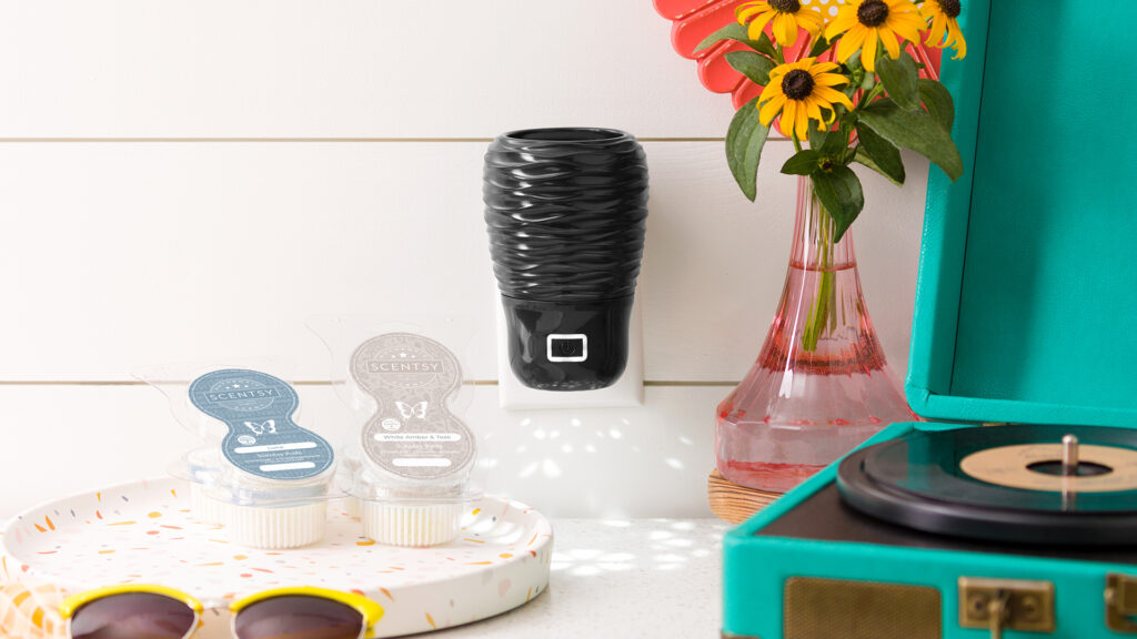 Scentsy wall fan diffuser and scentsy pods sitting on a counter next to a teal record player, pink vase of flowers, and yellow sunglasses