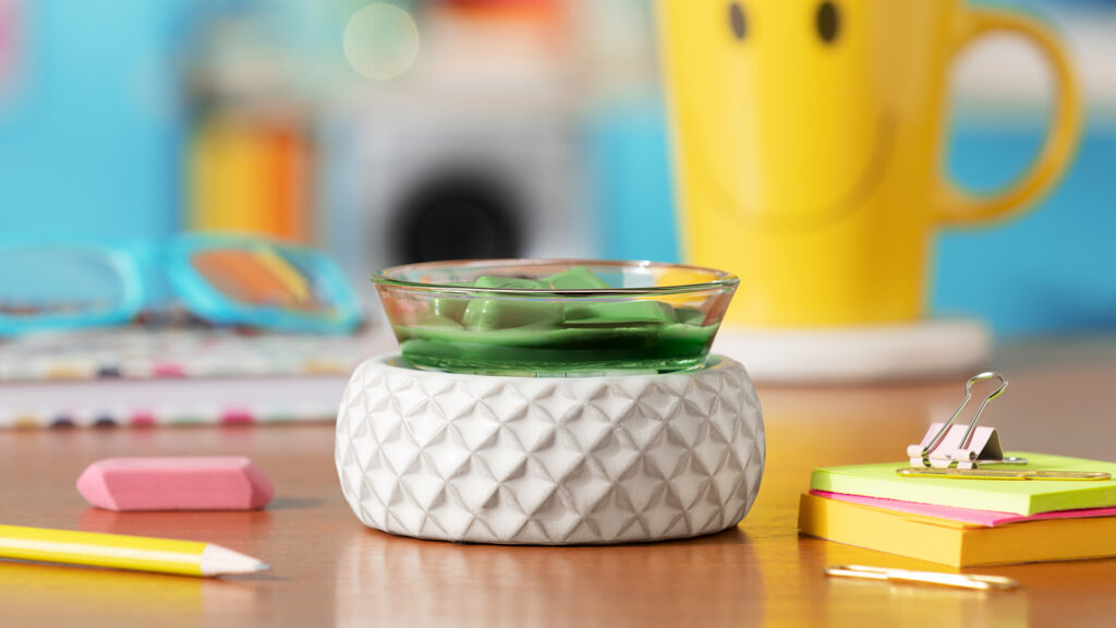 Scentsy wax warmer with green wax melts sitting on a desk with school supplies, glasses and a coffee mug 