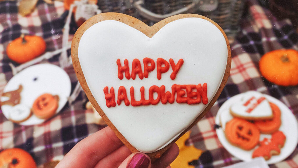 A woman's hand with red painted nails holding a heart shaped cookie frosted with white icing and happy halloween written in orange with a plaid table cloth and halloween decorated cookies in the background