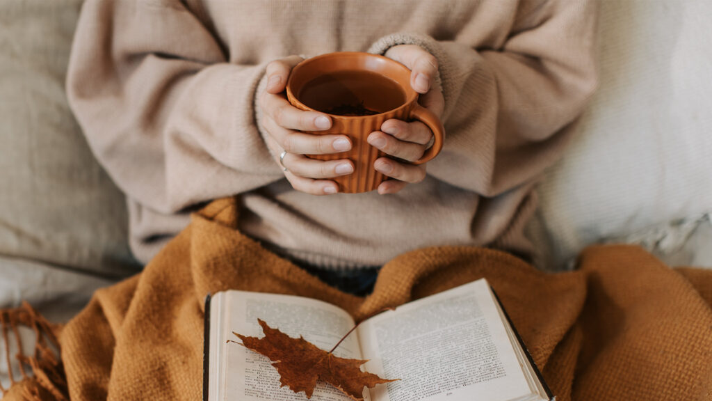 A girl sitting on a couch holding an orange cup of coffee with and orange blanket and book in her lap