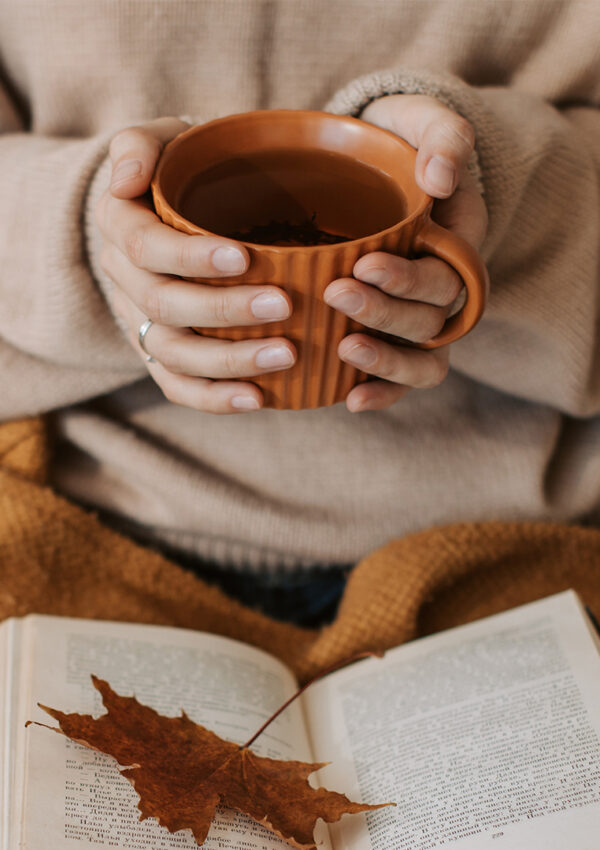 A girl sitting on a couch holding an orange cup of coffee with and orange blanket and book in her lap
