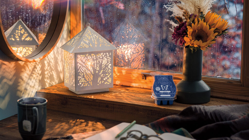 Forest light wax warmer lit up beside a rainy window, vase of flowers, cup of tea and blue wax melts