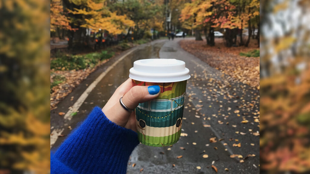 A woman's hand with blue painted nails holding up a coffee cup with a fall landscape in the background with a wet black road and orange and yellow falling leaves